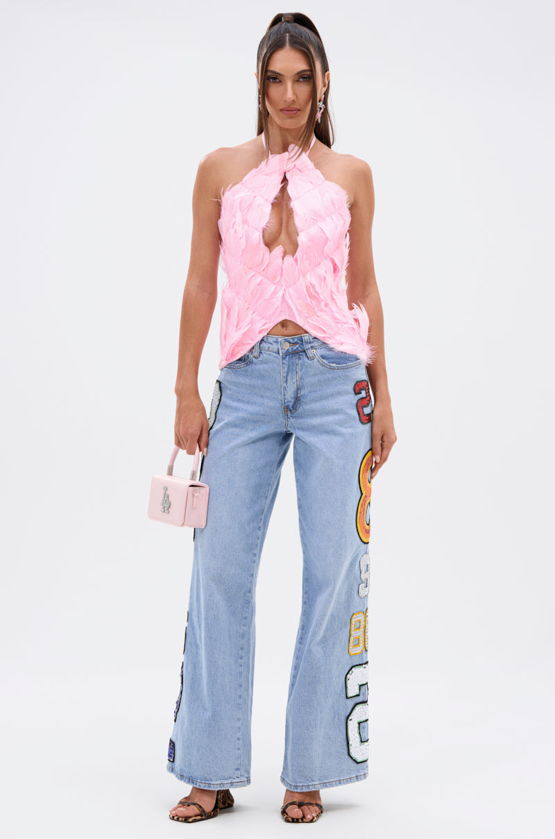 A woman stands confidently facing forward in the FEATHER QUEEN BLOUSE, a pink feather halter top with an open back, paired with light blue wide-leg jeans featuring colorful patches and numbers, strappy heels, and a small pink handbag. Her long hair completes the bold look.