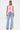 A woman stands confidently facing forward in the FEATHER QUEEN BLOUSE, a pink feather halter top with an open back, paired with light blue wide-leg jeans featuring colorful patches and numbers, strappy heels, and a small pink handbag. Her long hair completes the bold look.