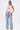 A woman models the FEATHER QUEEN BLOUSE in sleeveless pink with a halter neckline and open back, teamed with wide-leg light blue jeans featuring large colorful numbers. She accessorizes with a small pink handbag and heeled sandals.
