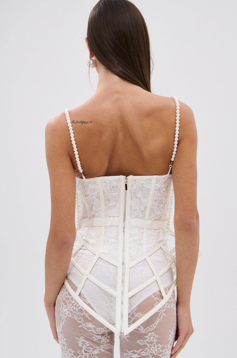 A woman faces away from the camera in the PEARL QUEEN CORSETED CAGE TOP—a white lace corset with a sweetheart neckline and faux pearl trim—paired with sheer lace pants. Her long hair is loose, revealing a small tattoo on her upper back.
