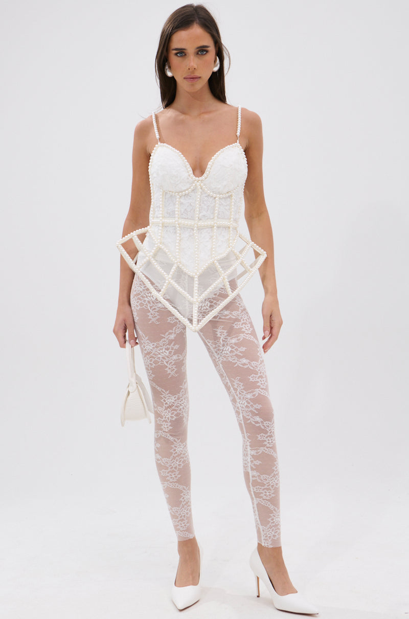 A woman poses against a white background wearing the PEARL QUEEN CORSETED CAGE TOP with a sweetheart neckline, sheer white lace leggings, white high heels, and holding a small white handbag.