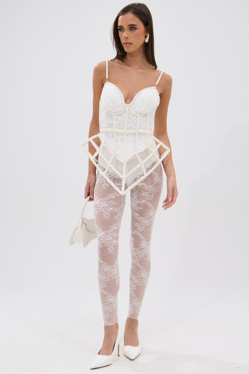 A woman wears the PEARL QUEEN CORSETED CAGE TOP with faux pearl trim, sheer lace leggings, white heels, statement earrings, and a white handbag while standing against a plain white background.