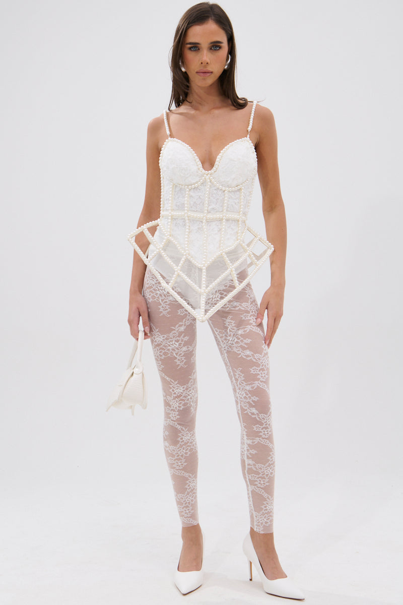 A woman poses against a white background wearing the PEARL QUEEN CORSETED CAGE TOP with geometric details and sweetheart neckline, paired with sheer lace leggings, white high heels, and a small white handbag.