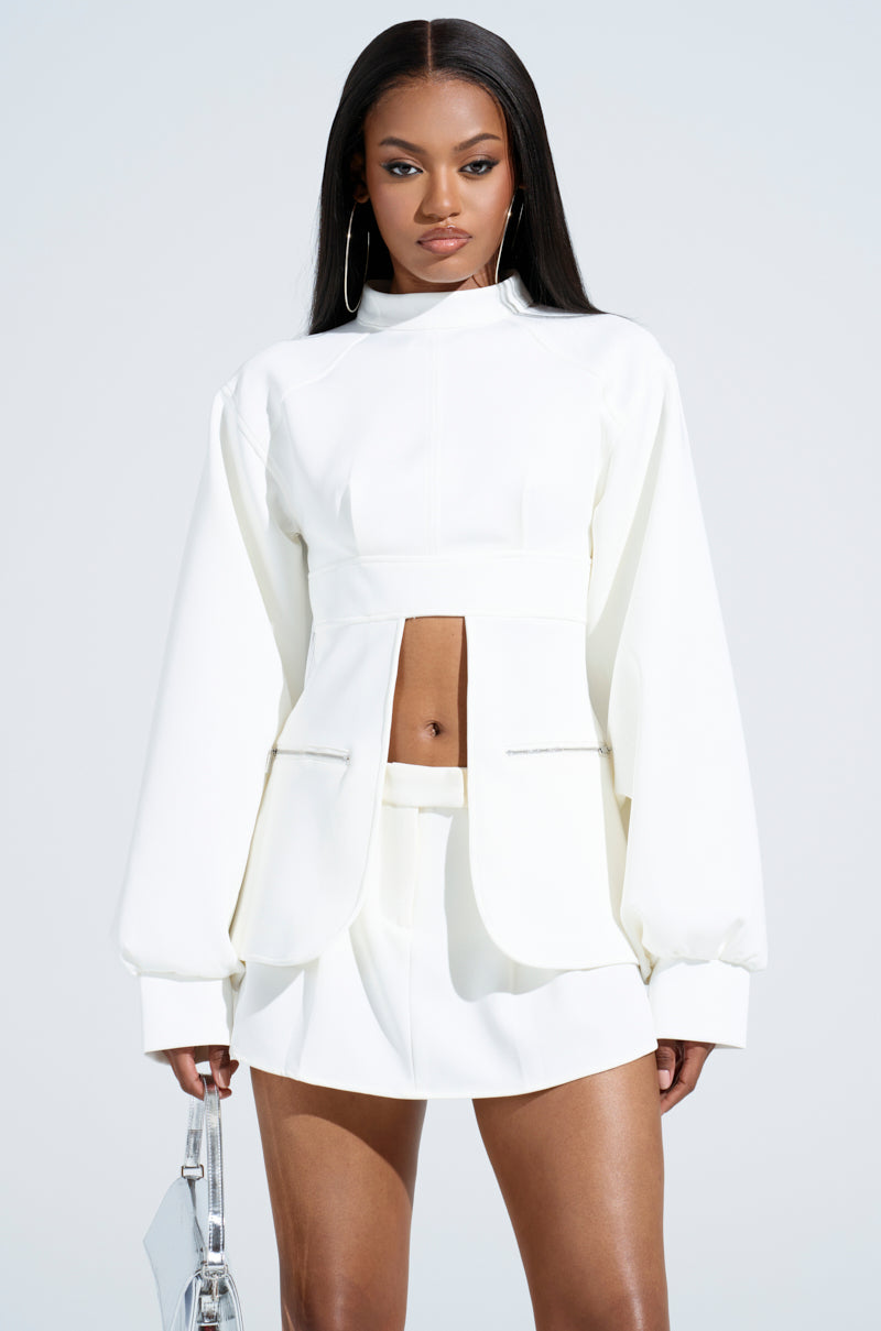 A woman stands confidently in a chic white outfit featuring the YOU WISH YOU WERE ME BLOUSE TOP with cut-out details and balloon cuffs, paired with a mini skirt. She holds a silver handbag and wears large hoop earrings, her straight and sleek hair contrasting against the soft white background.