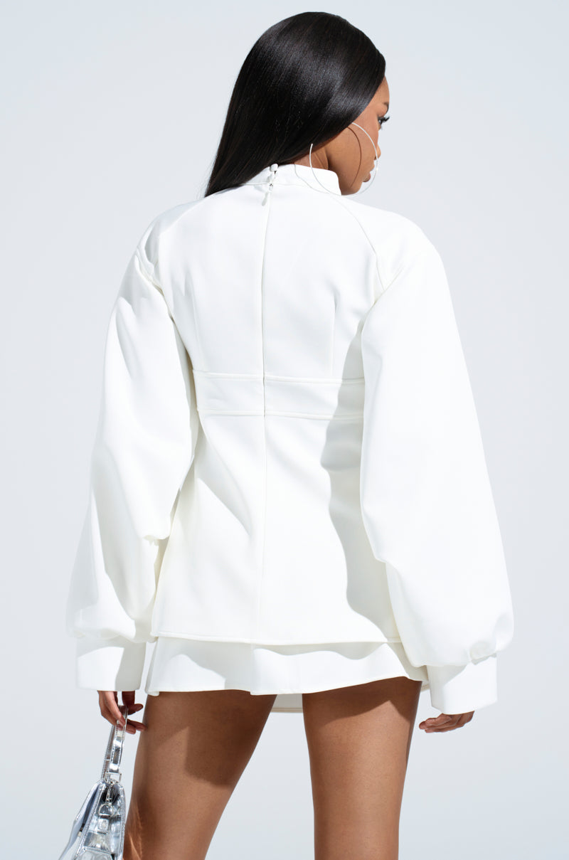 A woman with long dark hair is seen from behind in a stylish YOU WISH YOU WERE ME BLOUSE TOP with balloon cuffs and a short hem, holding a shiny silver handbag. Her large hoop earrings stand out against the plain light background.