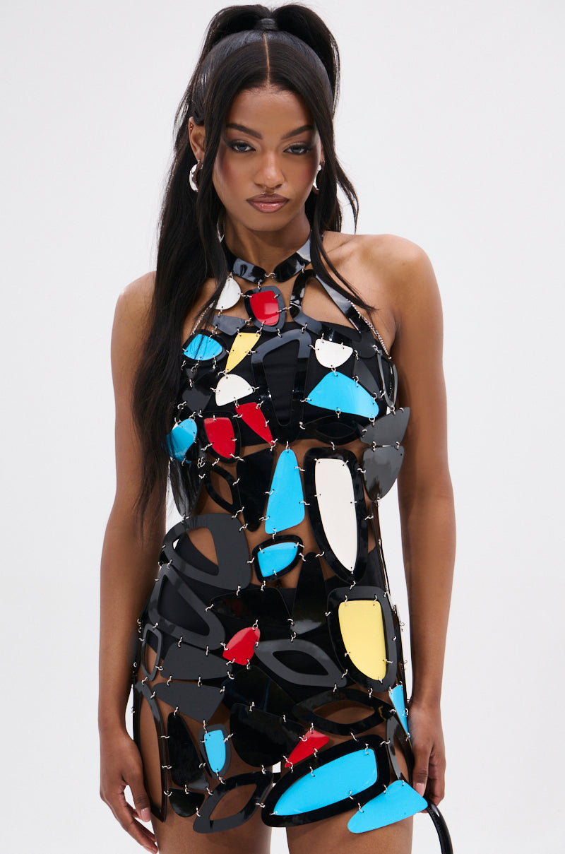 A person with long, dark hair wears the "A MASTERPIECE GEOMETRIC MINI DRESS," featuring interlocking abstract shapes in black, red, yellow, and blue. The vibrant design and halter neckline evoke Abstract Expressionism against a plain white background.