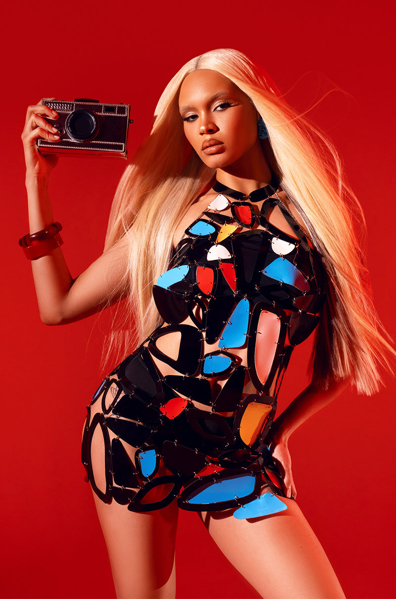 A woman with long blonde hair poses against a red background, holding a camera. She wears the A MASTERPIECE GEOMETRIC MINI DRESS, featuring black straps and bold red, blue, orange, and white geometric panels.