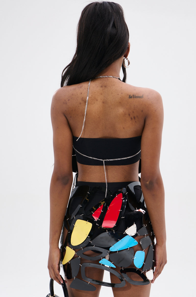 A person with long hair faces away from the camera, wearing a black top and the A MASTERPIECE GEOMETRIC MINI DRESS featuring colorful, irregular designs. A thin chain drapes over their shoulders, enhancing the ensemble's allure.