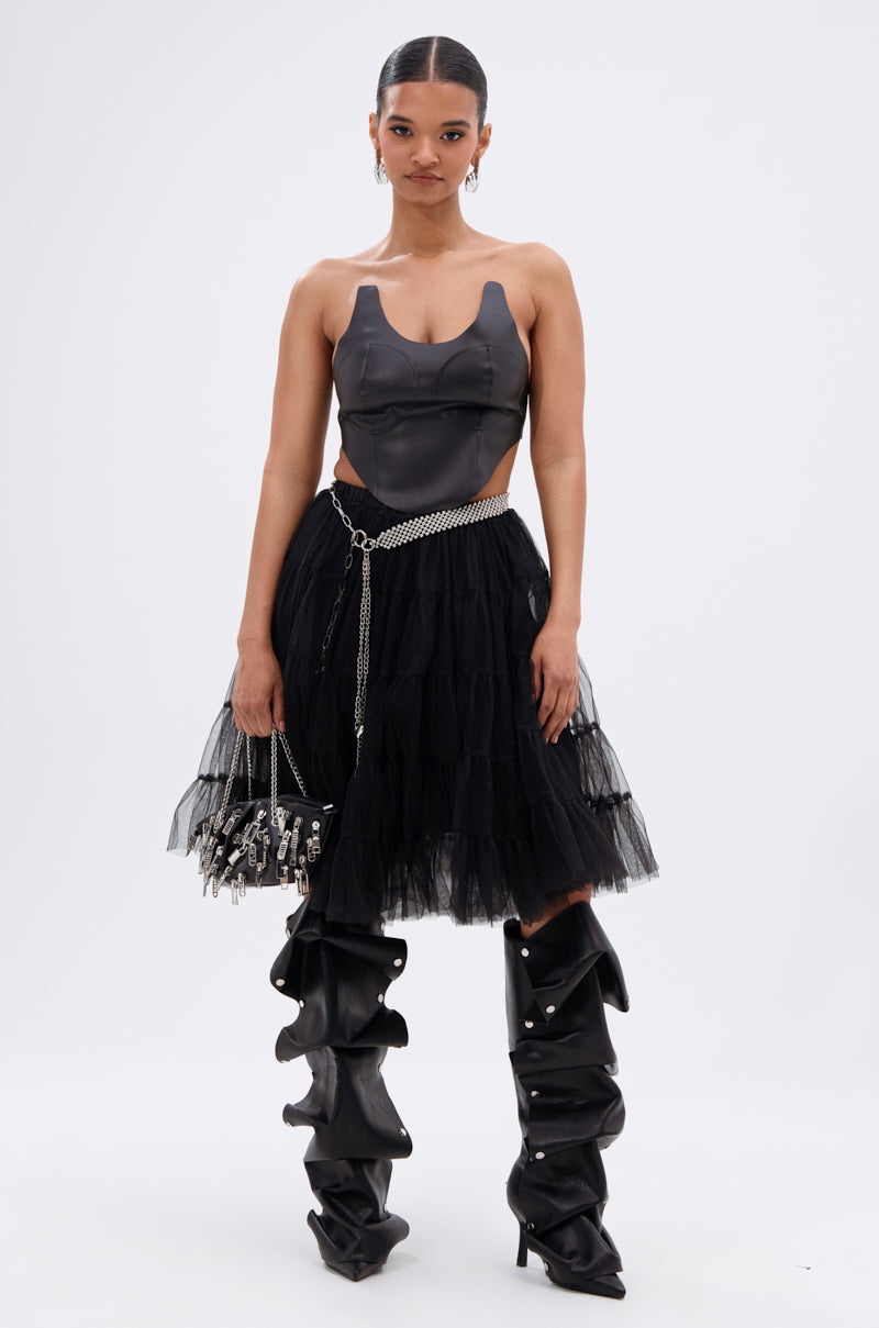 A woman faces forward in the SAMMI FAUX LEATHER CORSET, paired with a black tulle skirt, chain belt, small handbag, and tall slouchy black boots with metallic accents against a plain white background. 5