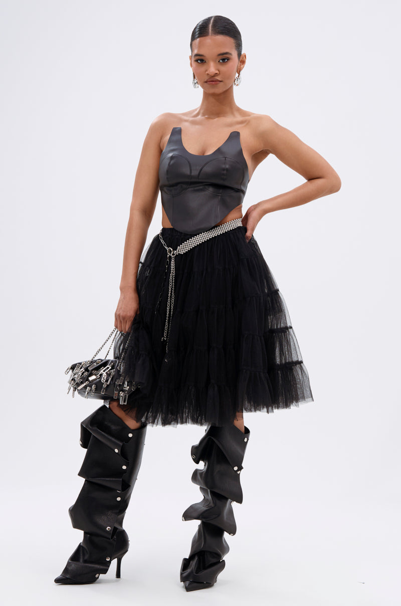 A woman poses confidently in the SAMMI FAUX LEATHER CORSET, paired with a layered black tulle skirt, slouchy high-heeled boots, a silver chain belt, and a small black handbag with silver details. 6