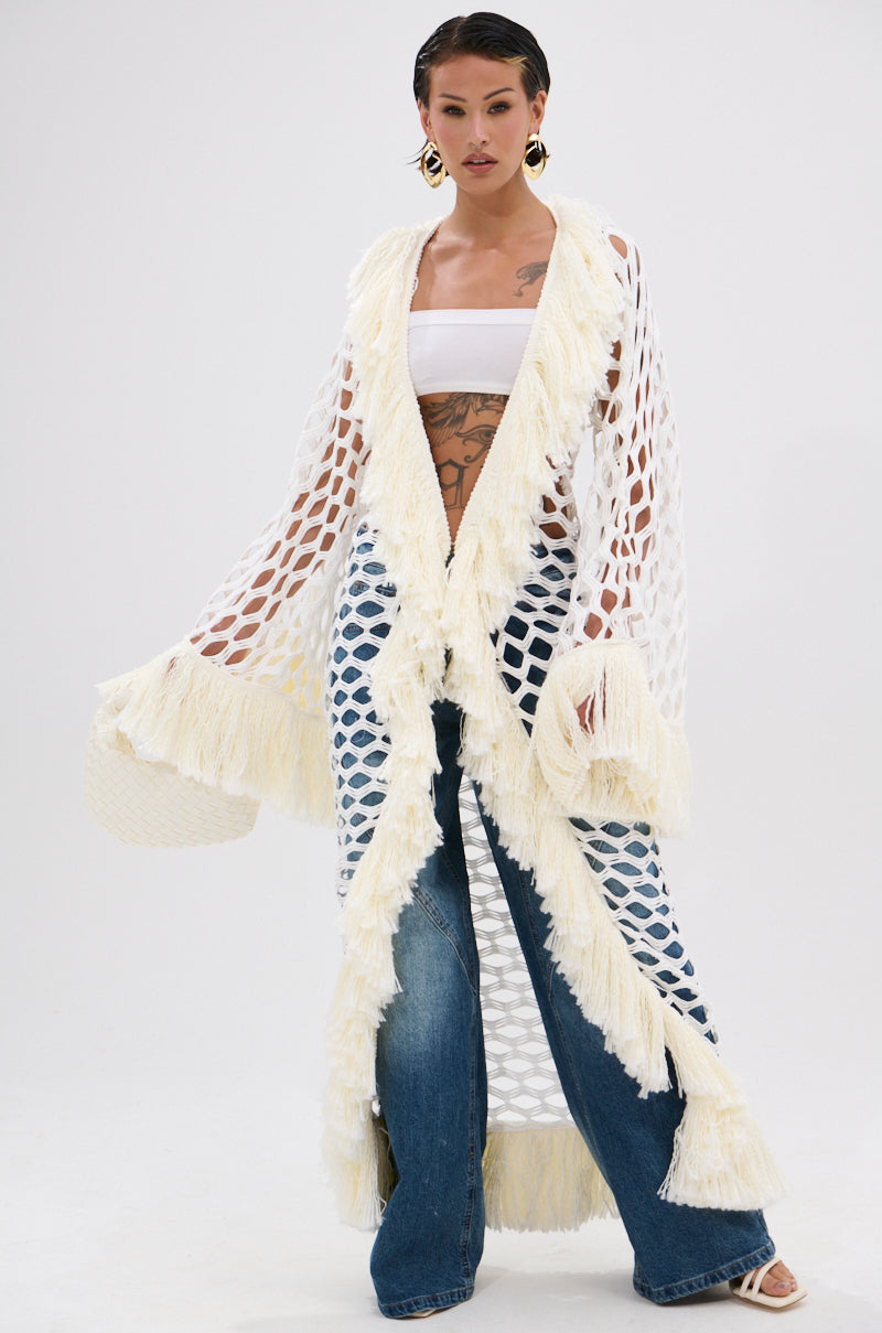 A woman models the BORN LIKE THIS FRINGE DETAIL LONG CARDIGAN, featuring a longline open-knit design with cream fringe accents, paired with a white bandeau top and wide-leg jeans against a plain white background.