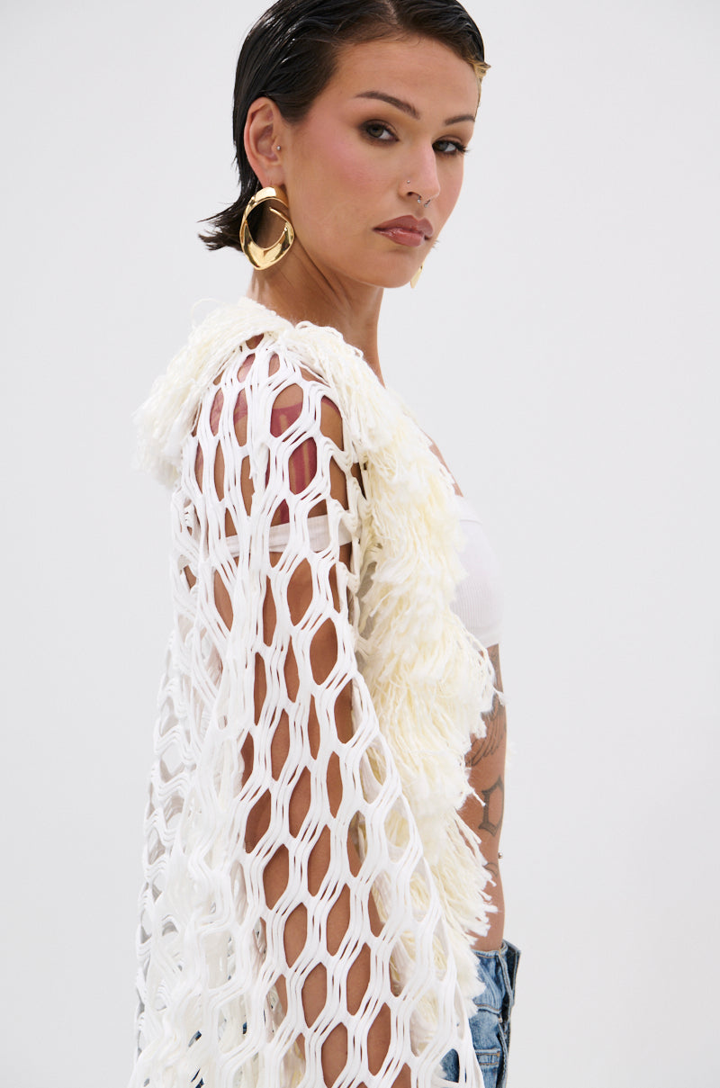 A person with short dark hair wears large gold hoop earrings and the BORN LIKE THIS FRINGE DETAIL LONG CARDIGAN over a white top, looking confidently at the camera against a plain background.