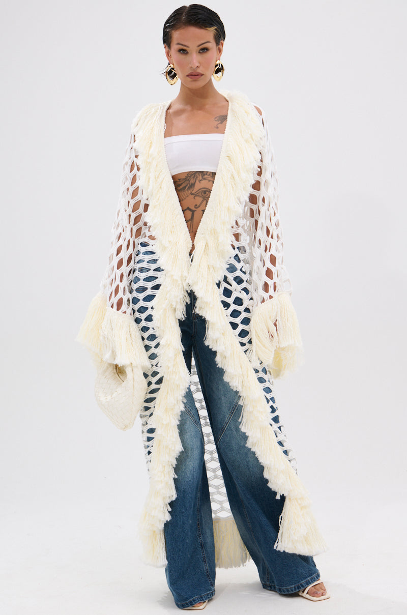 A woman models the BORN LIKE THIS FRINGE DETAIL LONG CARDIGAN, a longline open-knit style with cream fringe trim, layered over a white bandeau top, wide-leg jeans, and heeled sandals. She accessorizes with hoop earrings and a clutch.