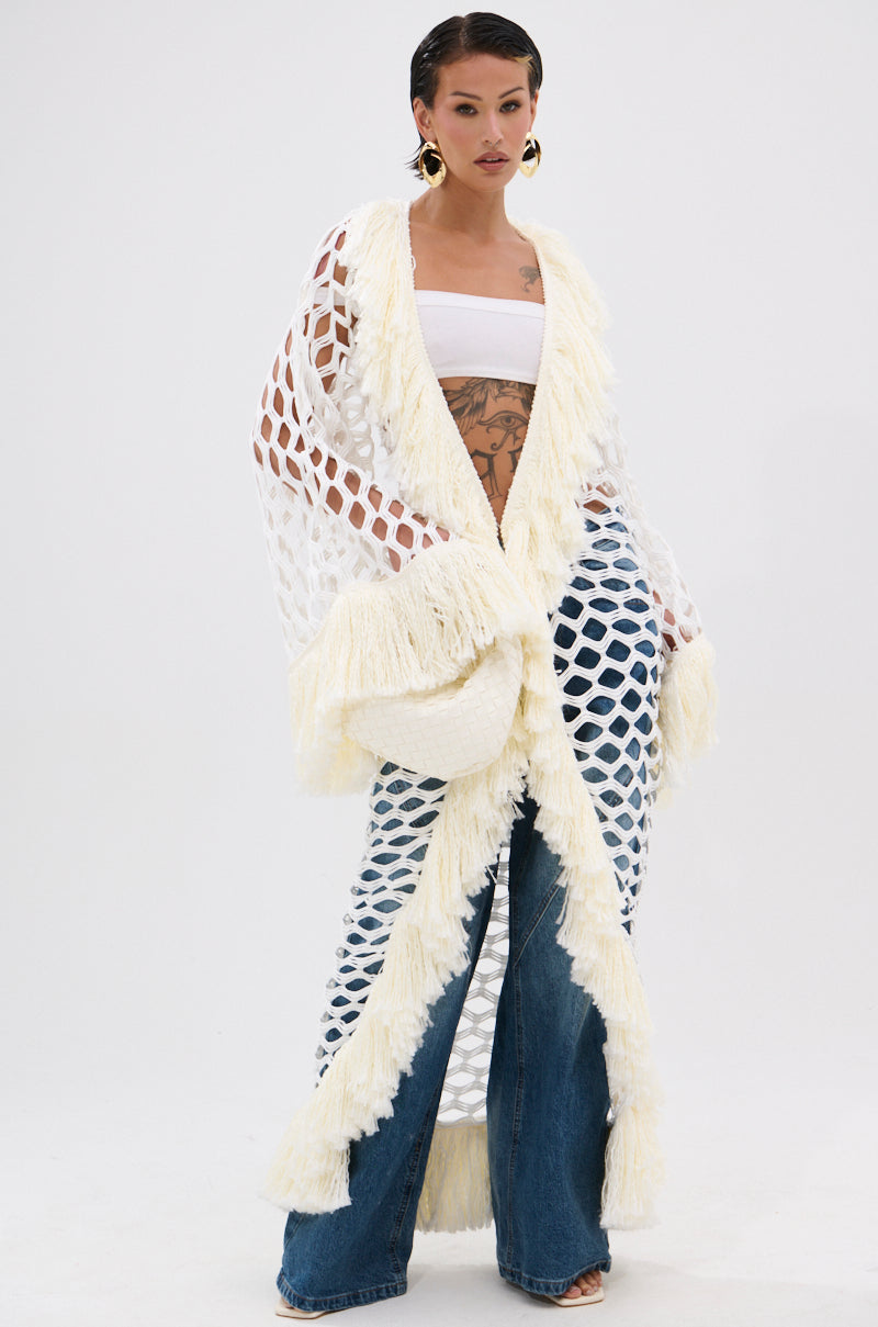 A woman poses in the BORN LIKE THIS FRINGE DETAIL LONG CARDIGAN over a white bandeau top and wide-leg blue jeans. She has short, slicked-back hair, large earrings, visible torso tattoos, and stands before a plain white background.