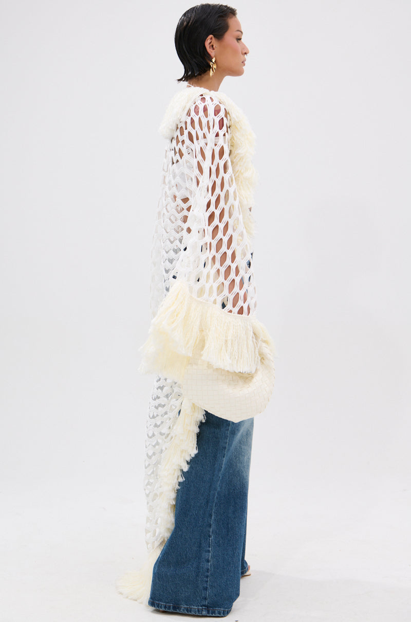 A person stands sideways in a longline silhouette, wearing the BORN LIKE THIS FRINGE DETAIL LONG CARDIGAN with open-knit large holes and fluffy fringe over blue wide-leg jeans. The background is plain white.