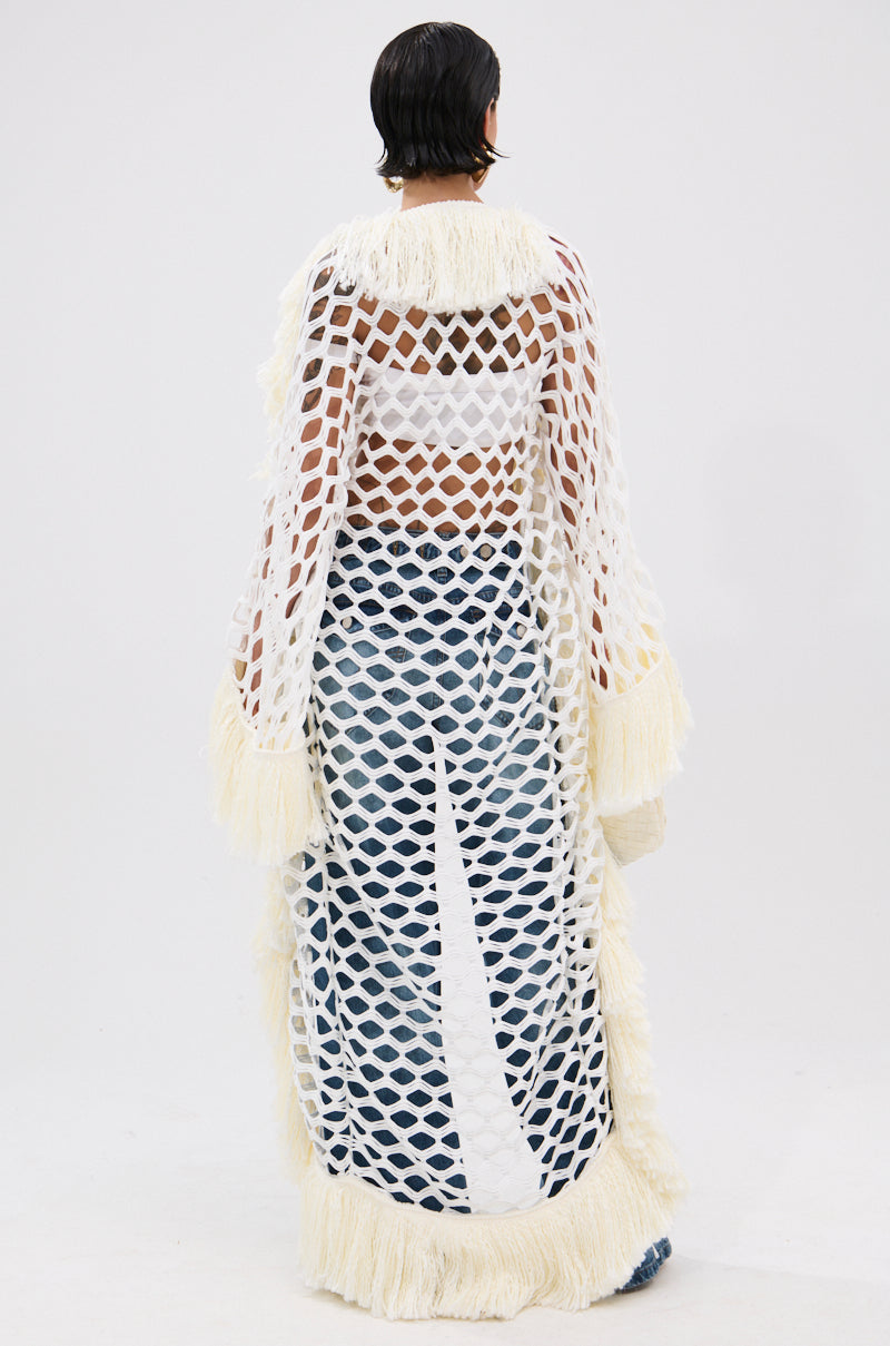 A person with short dark hair faces away, wearing the BORN LIKE THIS FRINGE DETAIL LONG CARDIGAN—an open-knit, longline white cardigan with fringe details—over blue pants and a white bralette. The background is plain white.