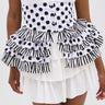 A person wears a white dress with polka dot and striped ruffled peplum corset, finished with the POLKA DOT IT BELT for a playful touch, featuring layered ruffles in both polka dot and striped patterns.