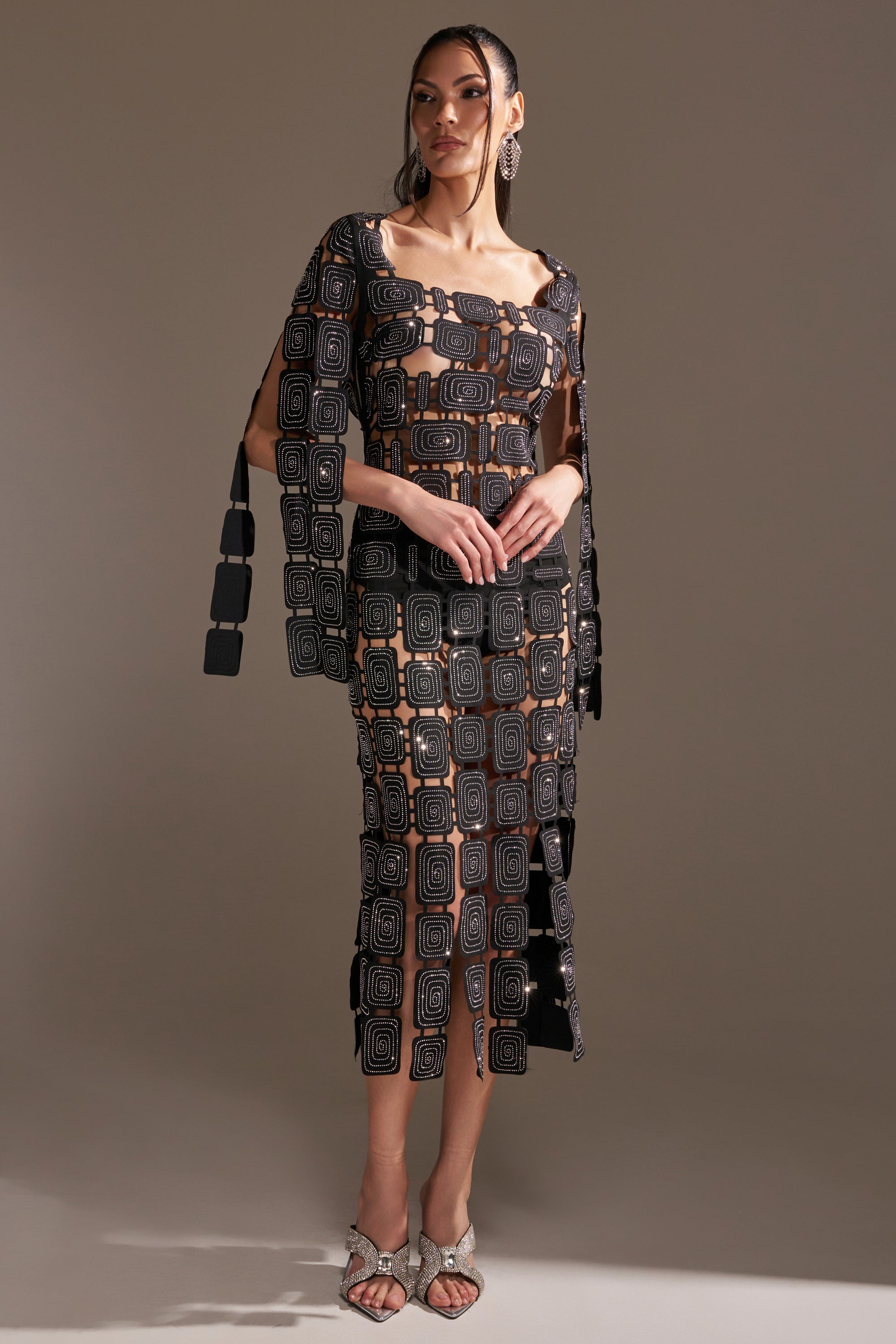 A woman stands in a studio wearing the ONLY ONE OF ME RHINESTONE MIDI DRESS, featuring geometric square patterns, a sheer bodycon silhouette, matching cape sleeves, and silver high-heeled sandals against a plain background.