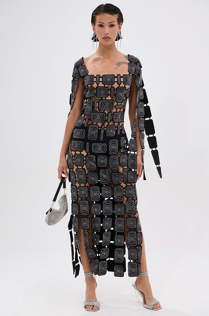 A woman poses in the ONLY ONE OF ME RHINESTONE MIDI DRESS—a sheer black bodycon midi with geometric square patterns and cutout details—paired with a silver handbag and silver high-heeled sandals against a white background.