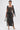 A woman poses in the ONLY ONE OF ME RHINESTONE MIDI DRESS—a sheer black bodycon midi with geometric square patterns and cutout details—paired with a silver handbag and silver high-heeled sandals against a white background.