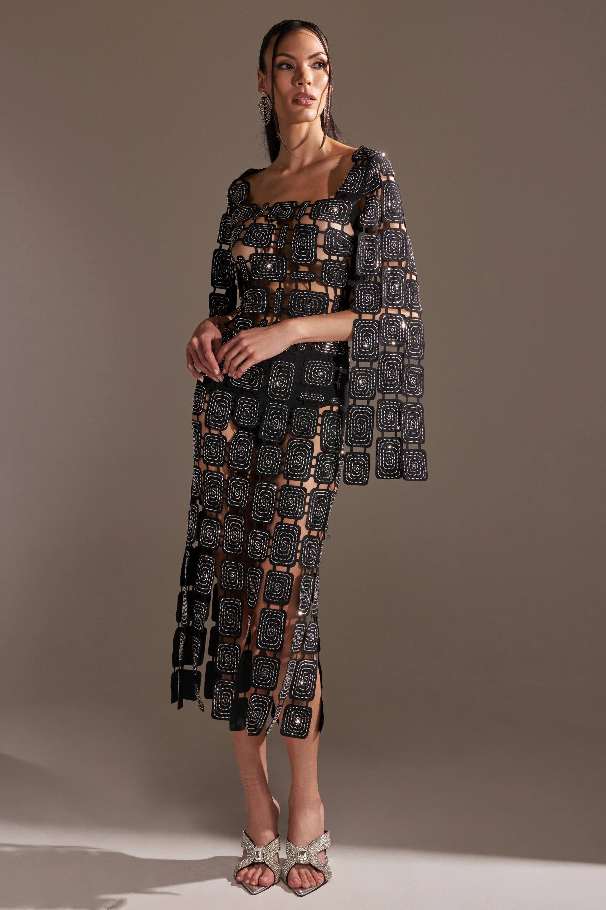 A woman poses in a studio, wearing the ONLY ONE OF ME RHINESTONE MIDI DRESS—a sheer, geometric-patterned black bodycon dress with square designs over a black undergarment—styled with statement earrings and open-toed heels against a neutral backdrop.