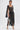 A woman poses against a white background in the ONLY ONE OF ME RHINESTONE MIDI DRESS, featuring sheer black fabric and bold geometric patterns. She styles it with silver heels, a small silver handbag, slicked-back hair, and statement earrings.