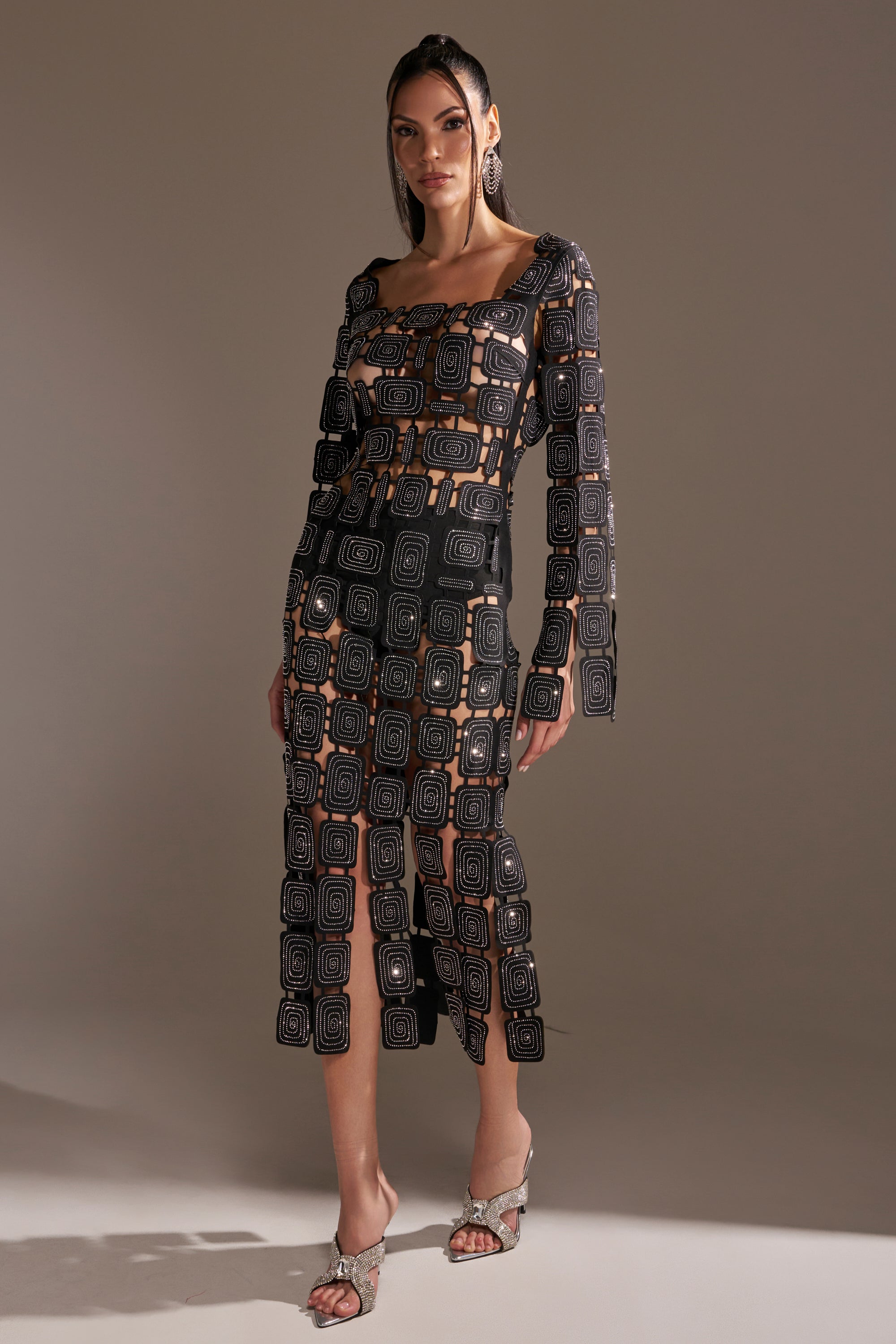 A woman models the ONLY ONE OF ME RHINESTONE MIDI DRESS—a sheer black, bodycon midi with geometric rhinestone patterns—paired with nude undergarments, long hair, large earrings, and open-toed heels in a neutral studio setting.