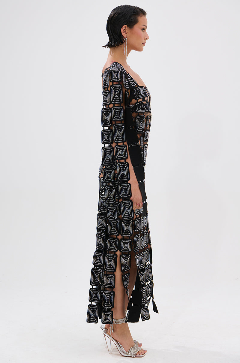 A woman with short dark hair stands in profile wearing the ONLY ONE OF ME RHINESTONE MIDI DRESS, a long black sheer bodycon dress with bold white geometric square patterns and high slits, paired with clear high heels against a plain white background.