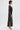 A woman with short dark hair stands in profile wearing the ONLY ONE OF ME RHINESTONE MIDI DRESS, a long black sheer bodycon dress with bold white geometric square patterns and high slits, paired with clear high heels against a plain white background.