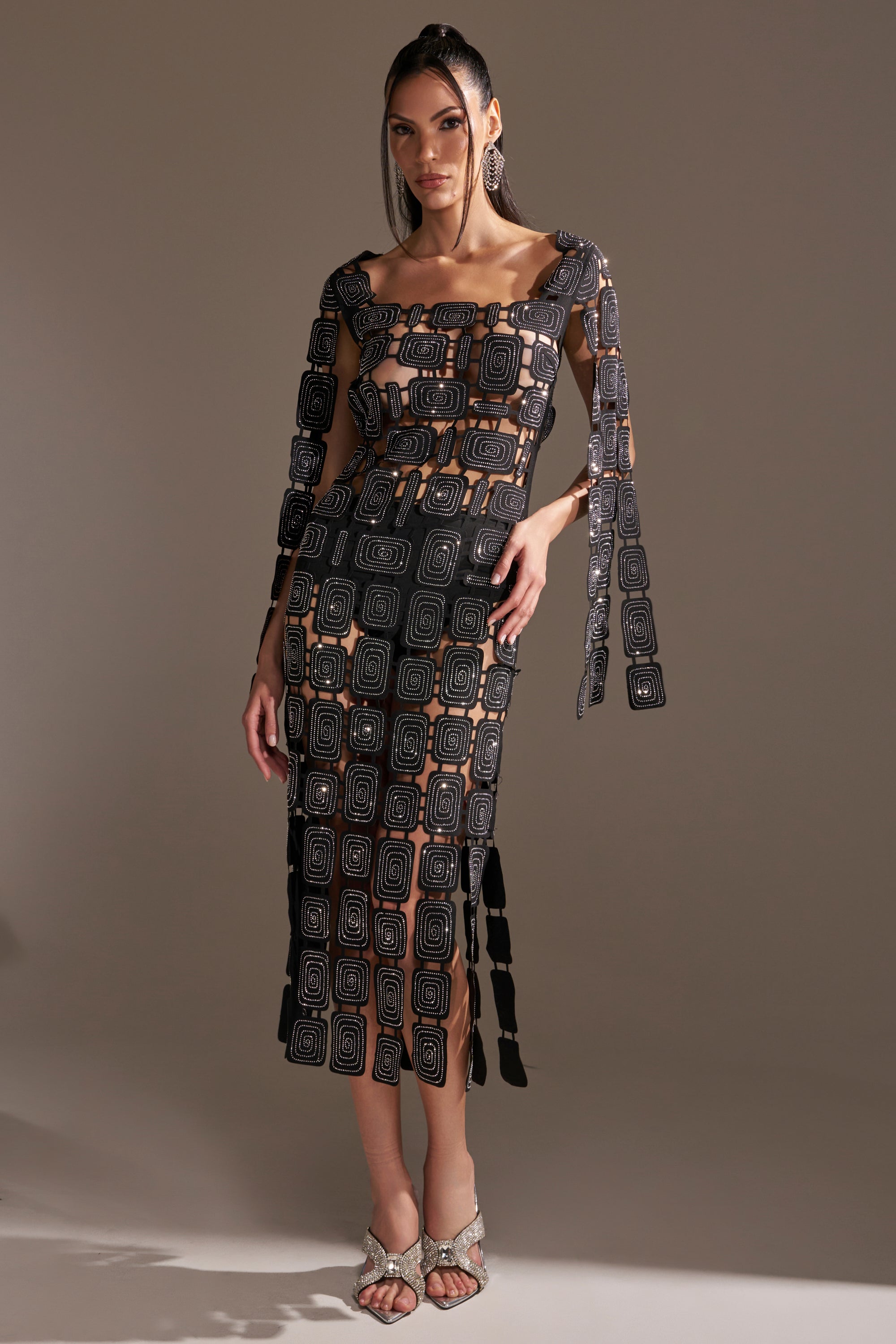 A woman stands in a studio wearing the ONLY ONE OF ME RHINESTONE MIDI DRESS, a black bodycon midi with geometric cutouts and wide shoulder straps. She accessorizes and completes her look with open-toed heels, her long hair styled loose.