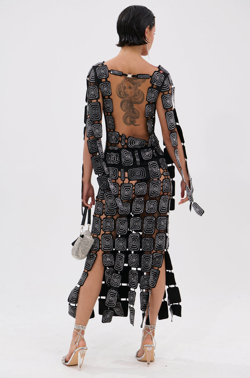 A person with short dark hair and a back tattoo wears the ONLY ONE OF ME RHINESTONE MIDI DRESS, featuring geometric square patterns and cutouts, paired with strappy heels and a sparkly handbag, against a plain white background.