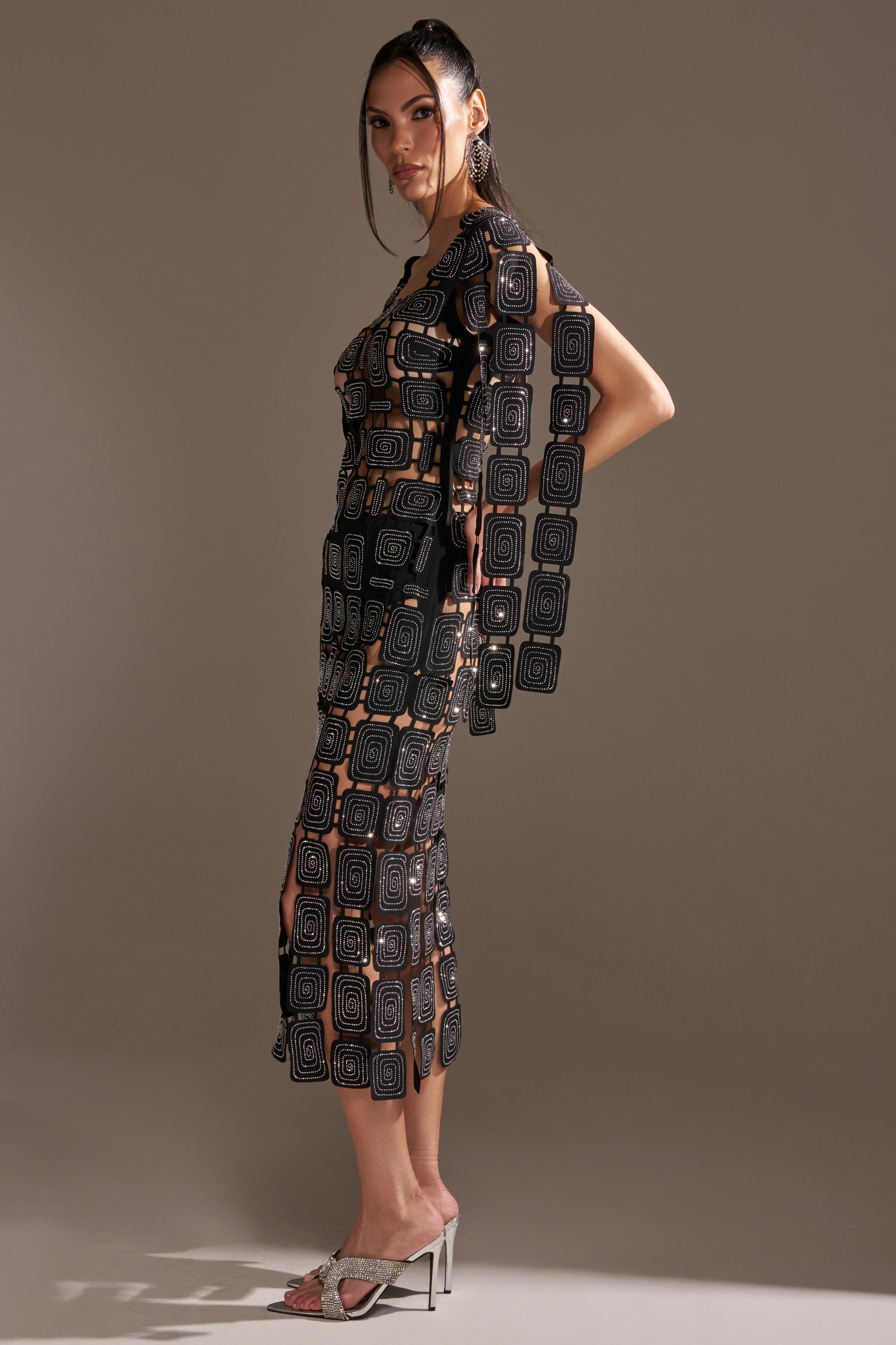 A woman stands in profile wearing the ONLY ONE OF ME RHINESTONE MIDI DRESS—a sheer black, geometric-patterned bodycon—paired with silver heels and statement earrings against a softly lit, plain background.