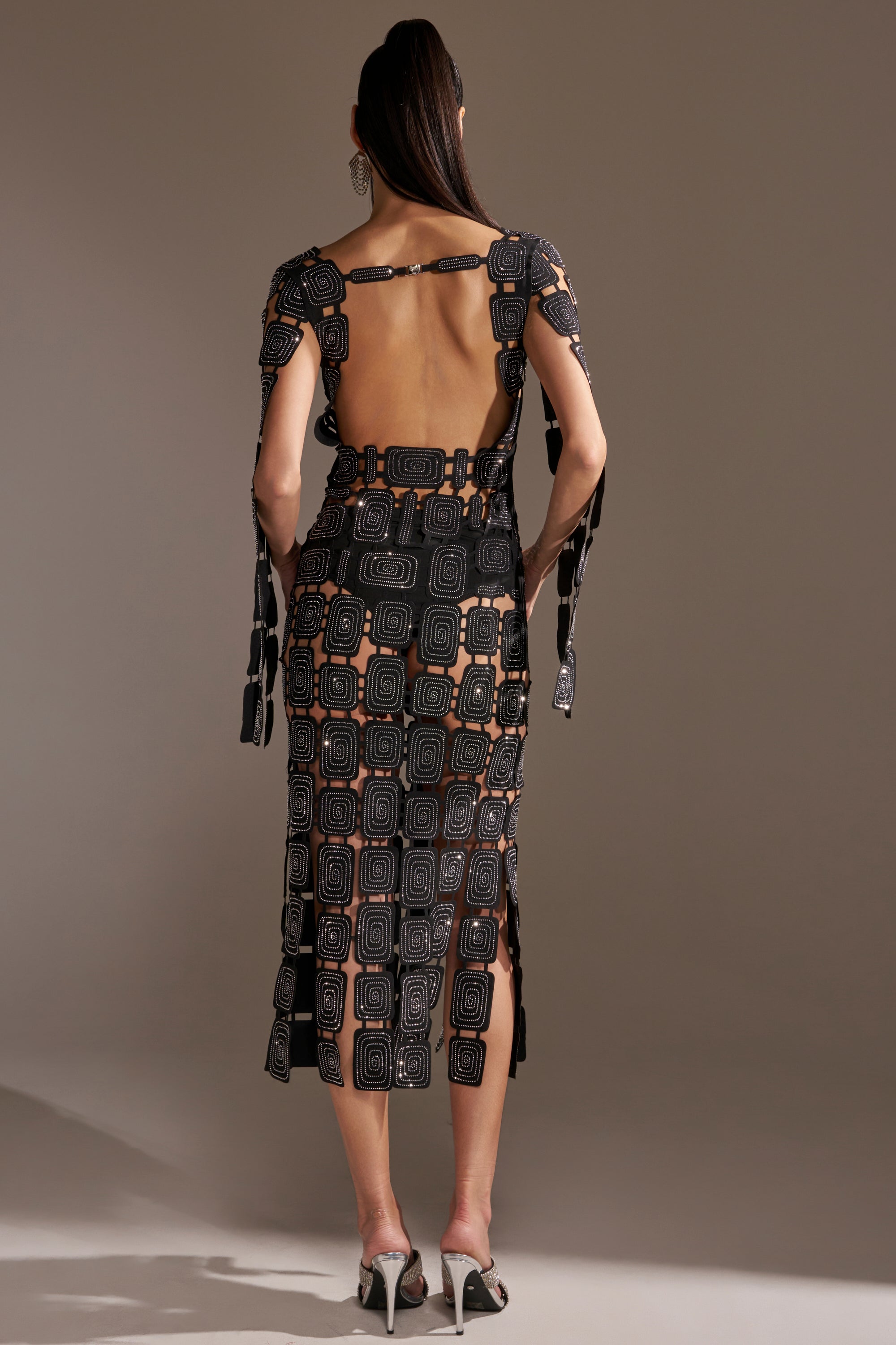 A woman with long dark hair tied back wears the ONLY ONE OF ME RHINESTONE MIDI DRESS, featuring black fabric with geometric rhinestone patterns, open back, side slits, strappy shoulder details, and white high heels.