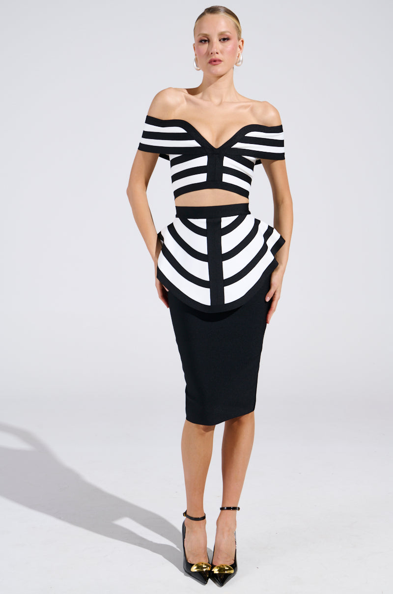A person models a black and white geometric off-shoulder top with a peplum overlay, paired with the WAITING FOR YOU MIDI SKIRT. Gold heels complement their neutral expression against a plain backdrop.