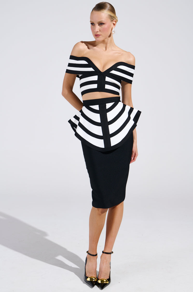 A woman confidently poses in the WAITING FOR YOU MIDI SKIRT featuring a black and white geometric off-shoulder design with peplum overlay. The bodycon silhouette is complemented by black heels with gold accents, all against a plain backdrop.