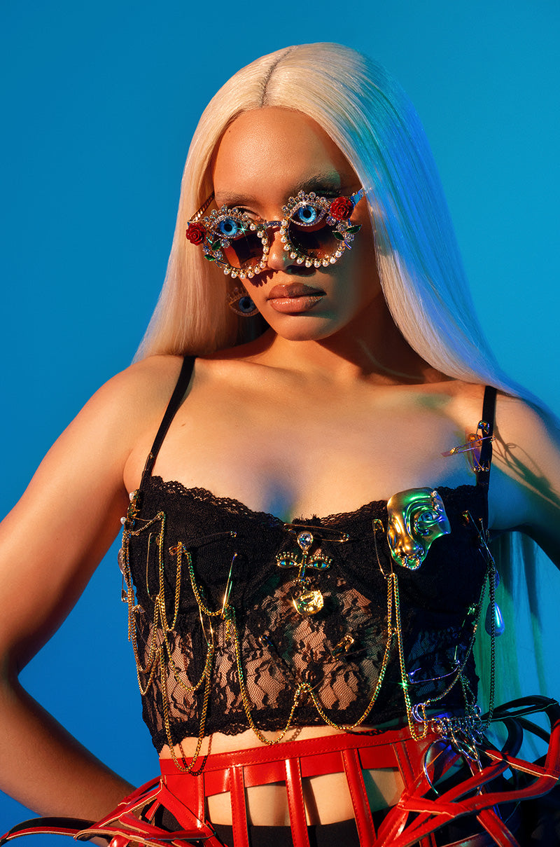 A person with long platinum blonde hair wears jeweled sunglasses, the TWO FACED PIN DETAILED LACE TOP with gold chains, and a red cage-like skirt, all set against a bright blue background.