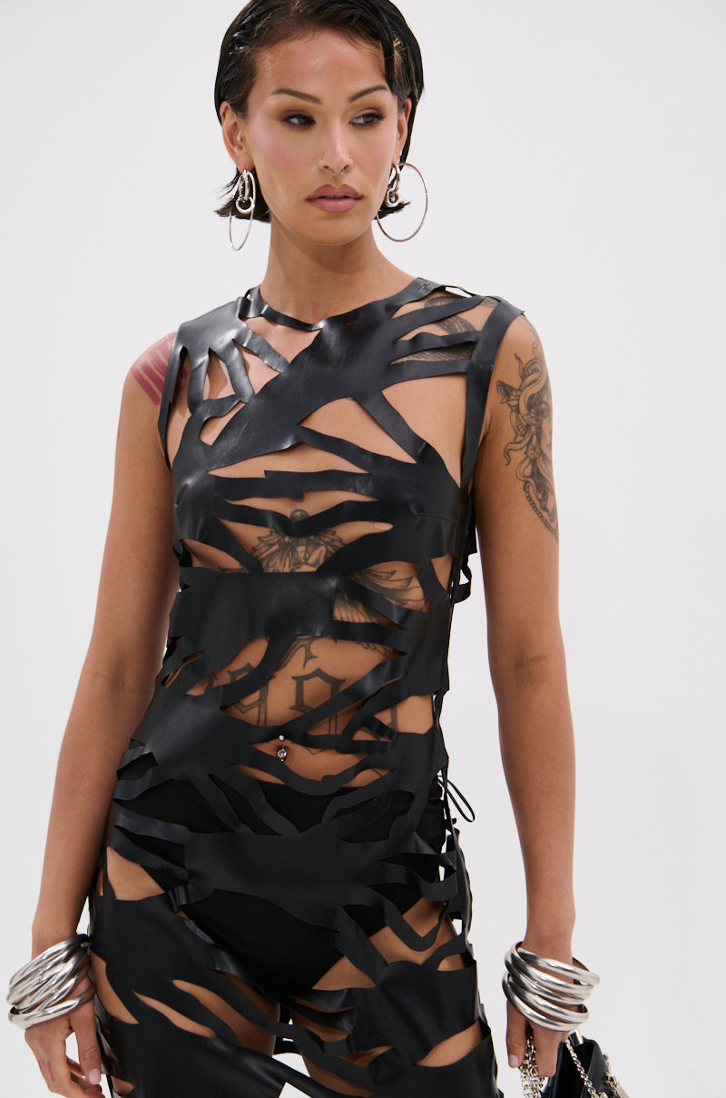 A woman wears the black ROUND OF APPLAUSE CUTOUT MAXI DRESS, revealing her black undergarments and tattoos. With short dark hair, hoop earrings, bold metal bracelets, and a bag in hand, she poses against a plain light background.
