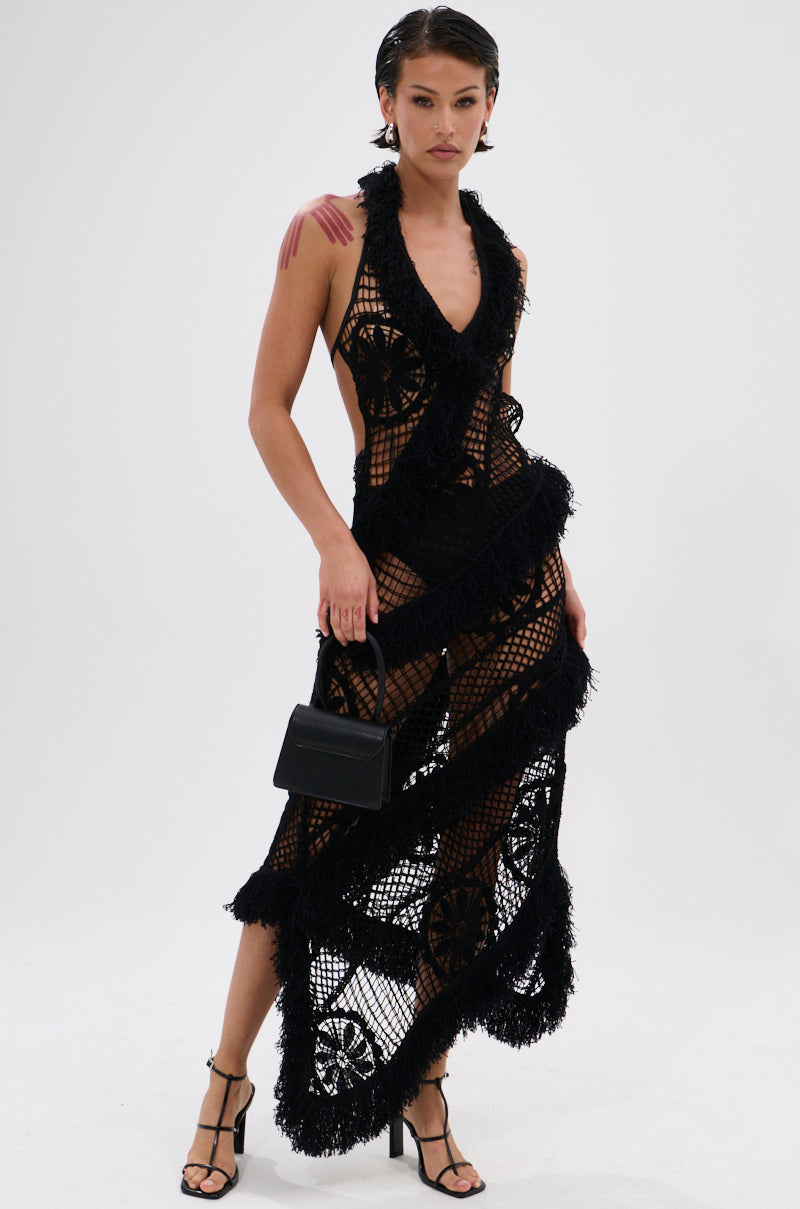 A woman poses in the MOMENTS IN TIME CROCHET MAXI DRESS, a sleeveless black maxi with fringe accents and a curve-hugging fit. She holds a small black handbag, wears strappy heels, and stands against a plain white background.