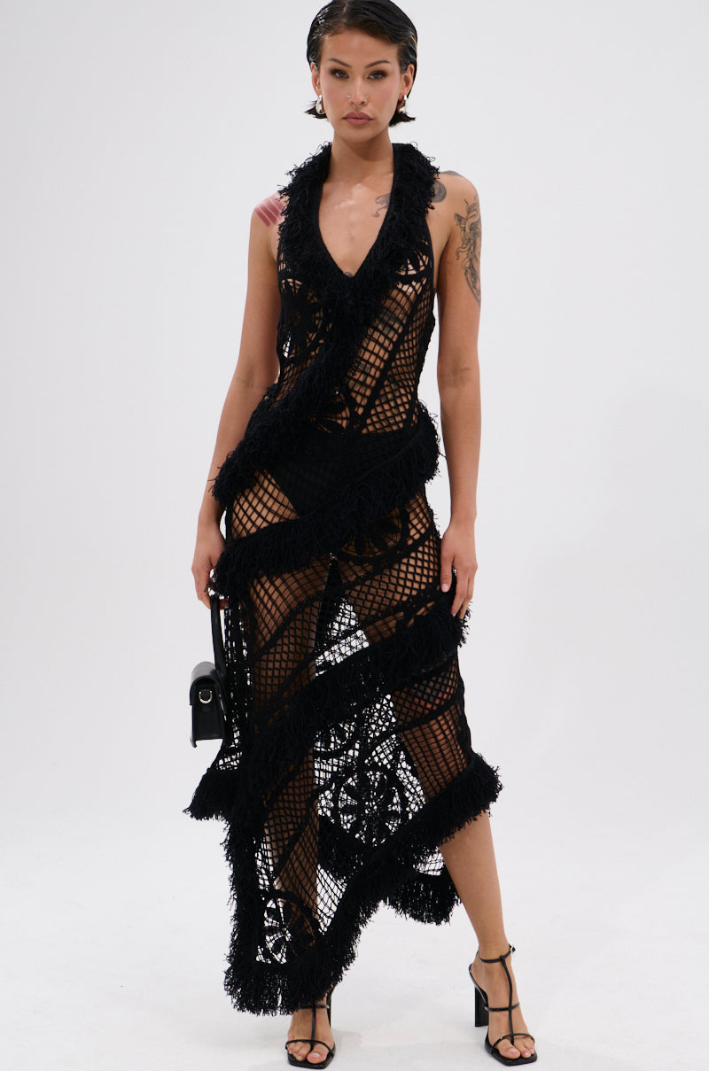 A woman with short hair and a tattooed arm poses against a white background in the MOMENTS IN TIME CROCHET MAXI DRESS—a black, sheer, netted maxi dress with textured stripes and an asymmetrical hem—paired with strappy heels and a small black handbag.