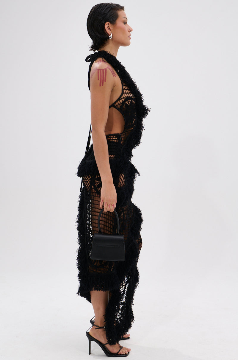 Against a plain white background, a model with slicked-back hair and visible arm tattoos wears the MOMENTS IN TIME CROCHET MAXI DRESS—a black, sheer, curve-hugging maxi over black undergarments, styled with high heels and a small black handbag.
