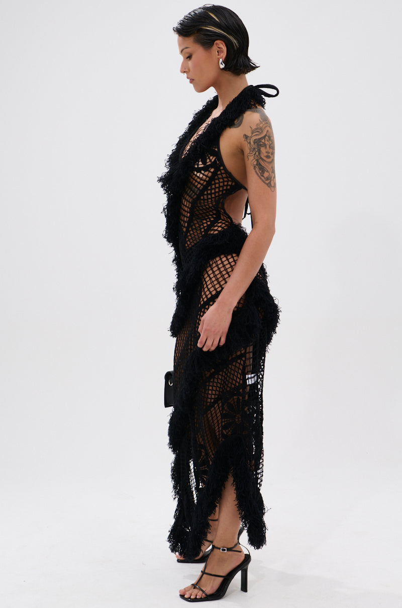 A person with short dark hair stands in profile against a white background, wearing the MOMENTS IN TIME CROCHET MAXI DRESS—black, fringed, see-through, and curve-hugging—over black undergarments and heeled sandals, showing a large arm tattoo.
