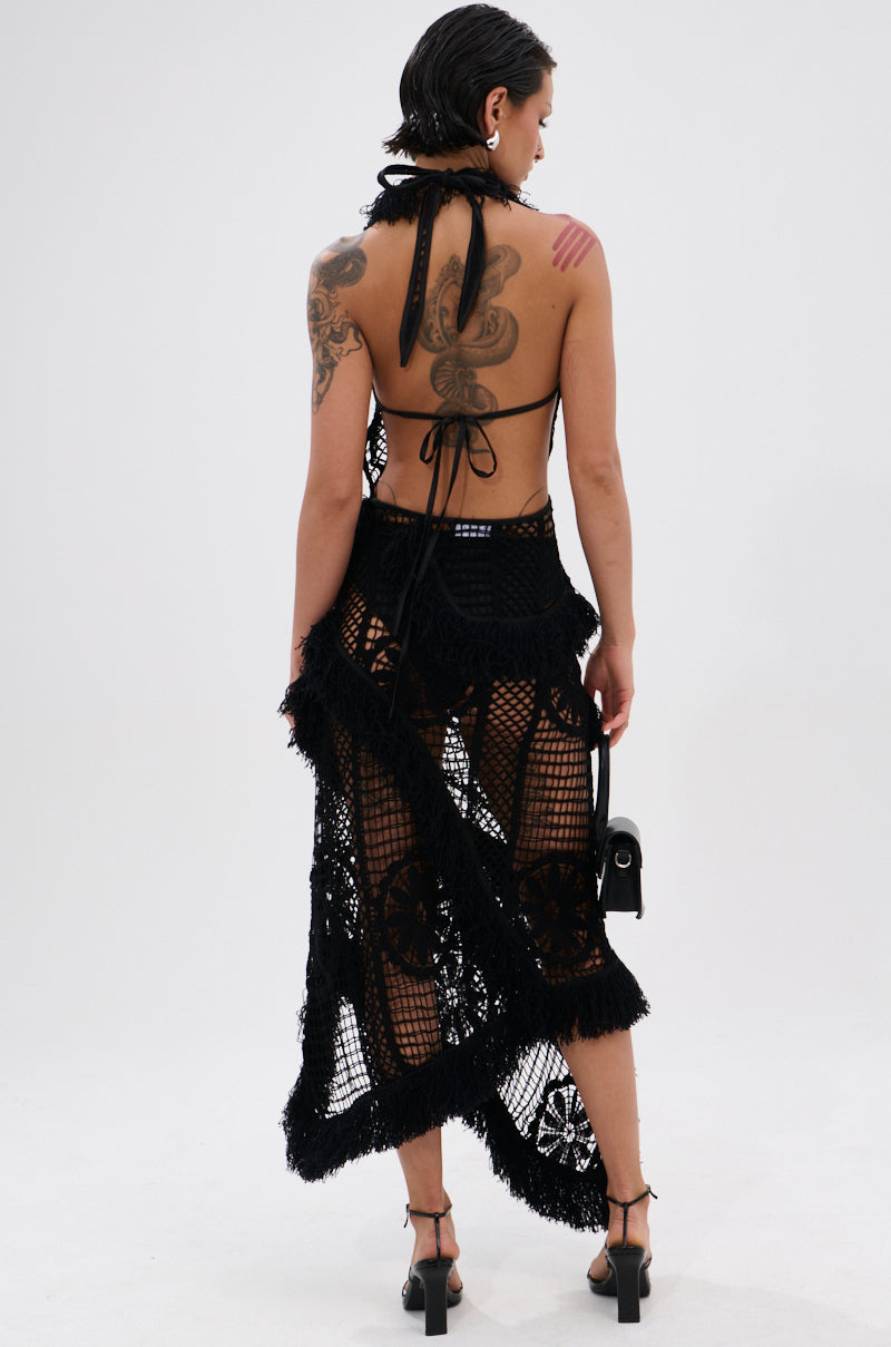 A woman with short dark hair, tattoos, and hoop earrings faces away in the MOMENTS IN TIME CROCHET MAXI DRESS—a black, curve-hugging crochet maxi—paired with black heels and a small black purse against a plain white background.