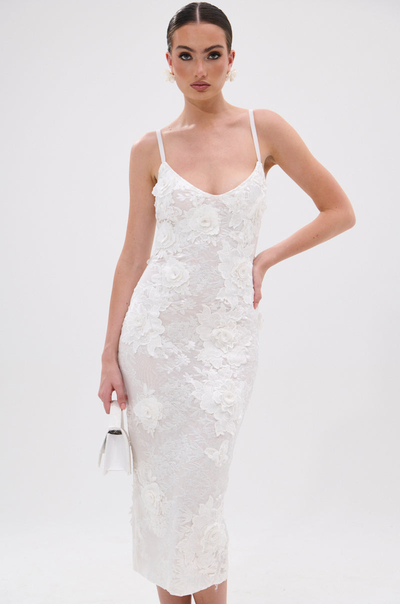 A woman in the IRIS FLORAL MIDI DRESS—a white lace midi with 3D floral appliqués, thin straps, and a bodycon fit—holds a small white handbag against a plain white background. 0