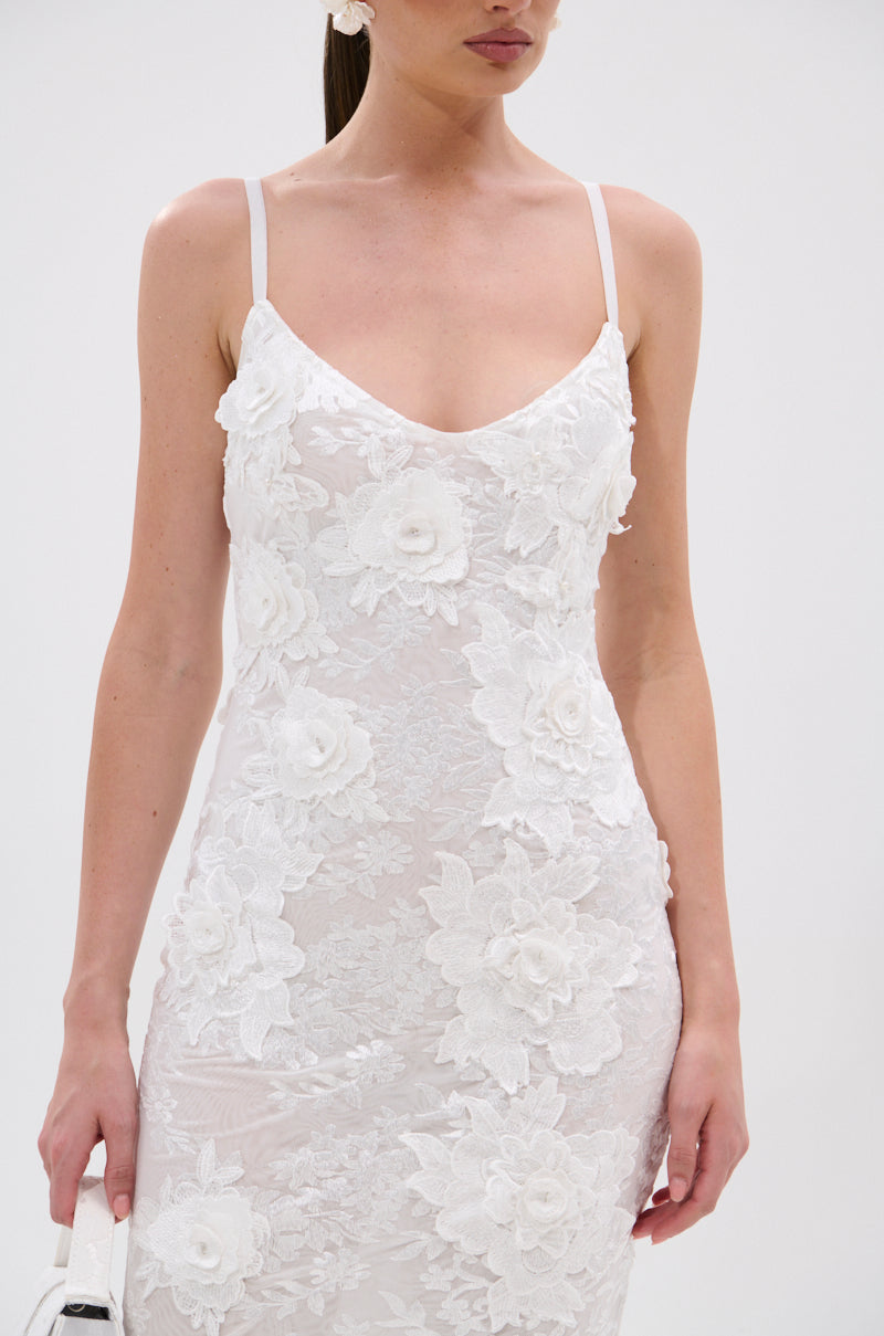 A woman models the IRIS FLORAL MIDI DRESS—a white, sleeveless bodycon dress with 3D floral appliqués—holding a small white bag, photographed from shoulders to mid-thigh against a plain background. 9