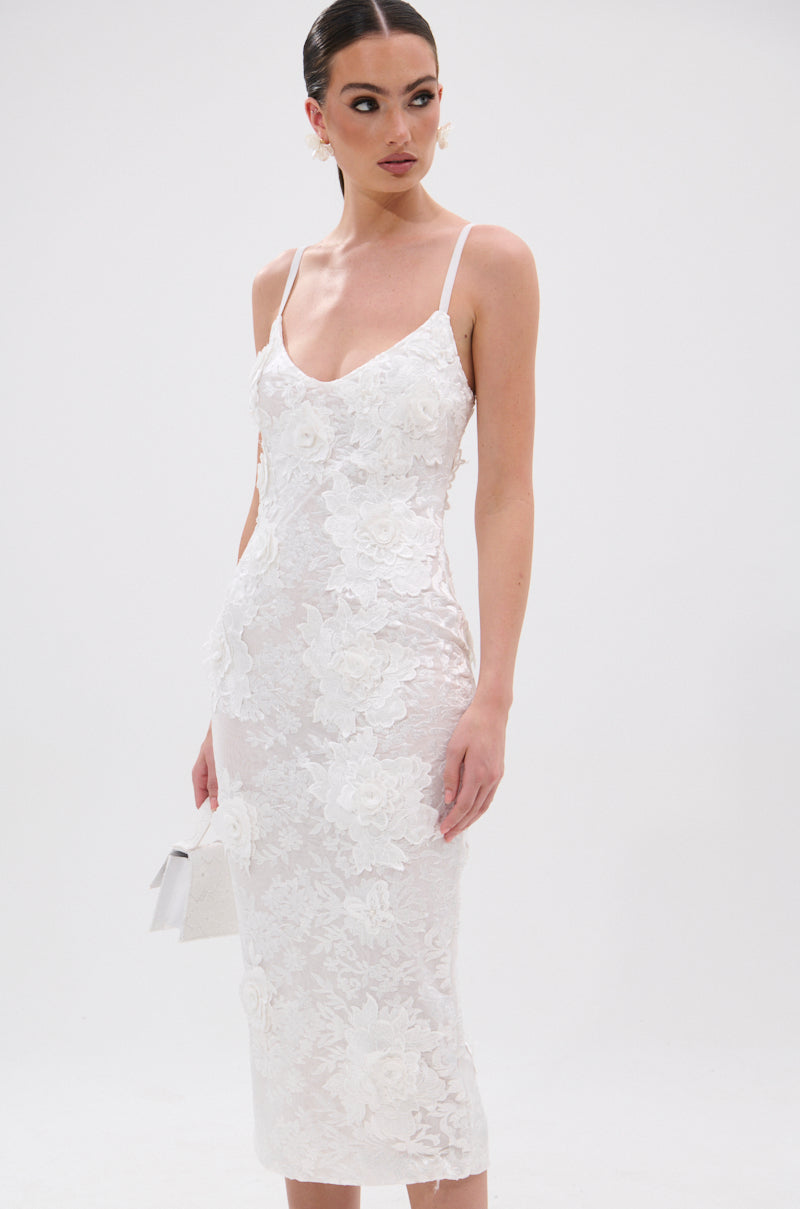 A woman in the IRIS FLORAL MIDI DRESS—a fitted white lace dress with floral patterns, thin straps, and a V-neckline—poses against a plain white background while holding a small white handbag. 1
