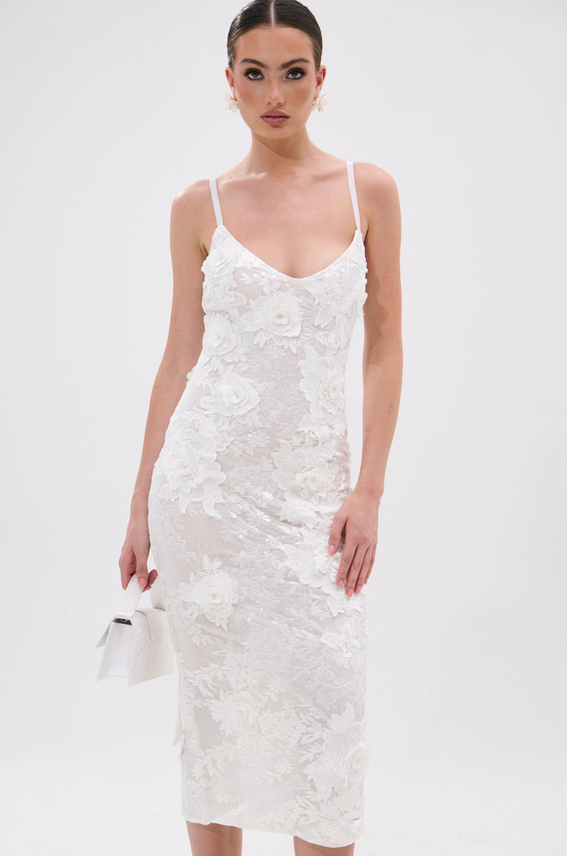 A woman in the IRIS FLORAL MIDI DRESS, featuring a bodycon fit and thin straps, holds a small white clutch while standing against a plain white background. 2