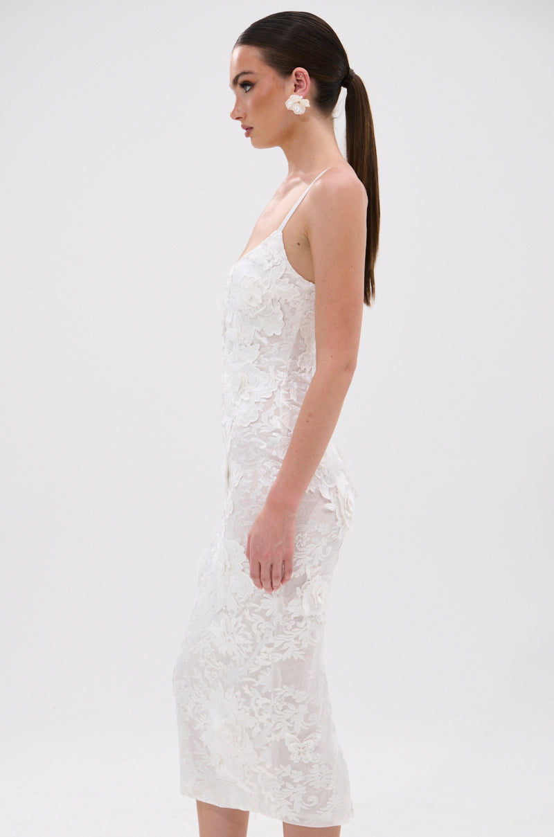 Woman in profile wears the IRIS FLORAL MIDI DRESS with 3D floral appliqués and thin straps, styled with large white floral earrings and long brown hair tied in a low ponytail, standing against a plain white background. 3