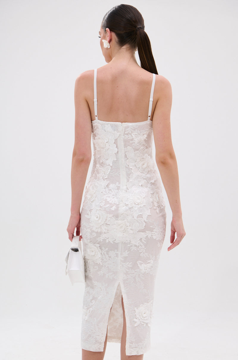 A woman with long brown hair in a low ponytail models the IRIS FLORAL MIDI DRESS, a bodycon, sleeveless dress with thin straps and 3D floral appliqués. She carries a small white handbag against a plain white background. 4
