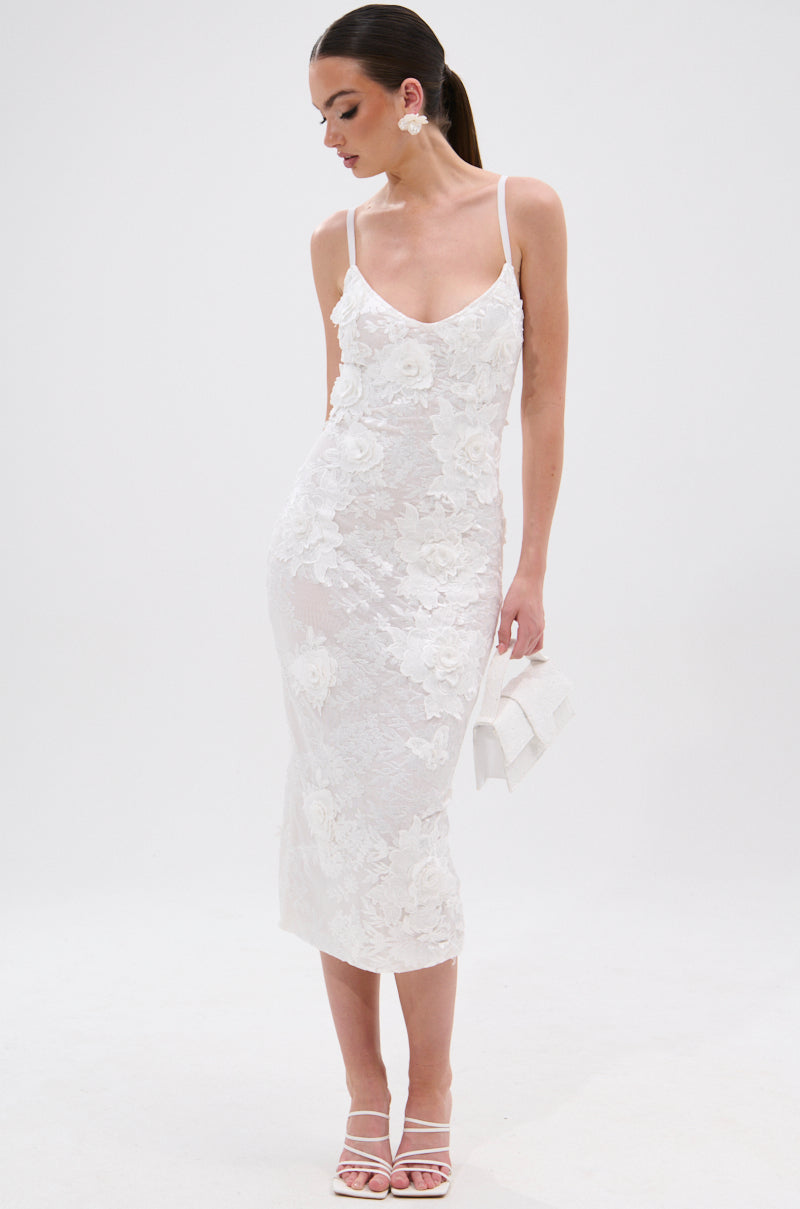 A woman in the IRIS FLORAL MIDI DRESS with a bodycon silhouette and 3D floral appliqués, white heels, statement earrings, and a white clutch stands against a plain white background. 5
