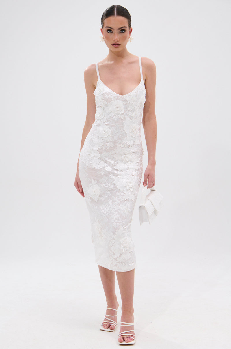 A woman in the IRIS FLORAL MIDI DRESS, with a fitted bodycon silhouette and thin straps, stands against a plain white background wearing white heeled sandals and holding a small white clutch. 6